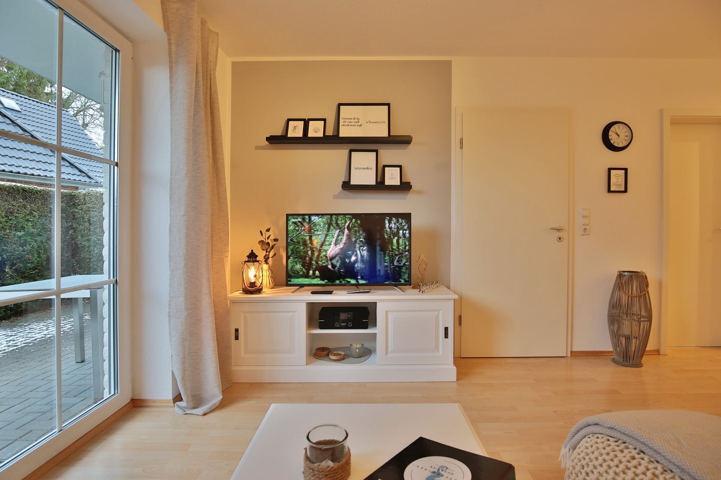 Ein Wohnzimmer mit Fernseher, einem Regal mit gerahmten Fotos, einer Uhr und einem Fenster mit Blick auf Bäume.
