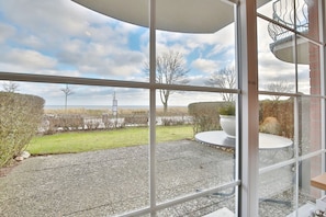 Une vue depuis une fenêtre donnant sur une terrasse en gravier, une table ronde et un pot de fleurs.