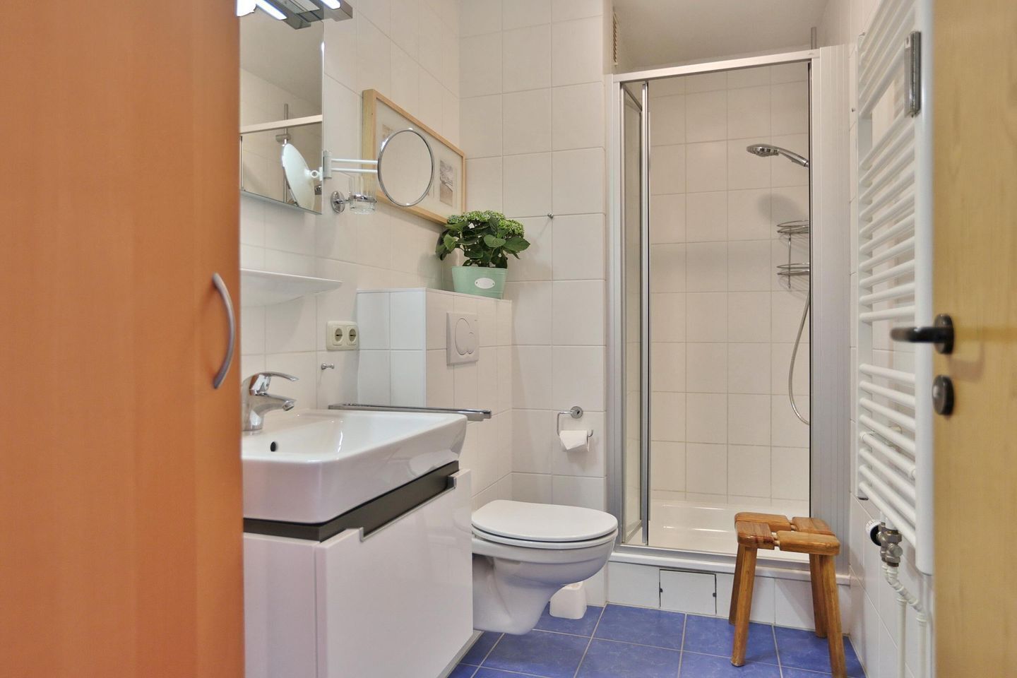 Baño con lavamanos, inodoro y ducha blancos. Se observa un taburete de madera y una planta en maceta.