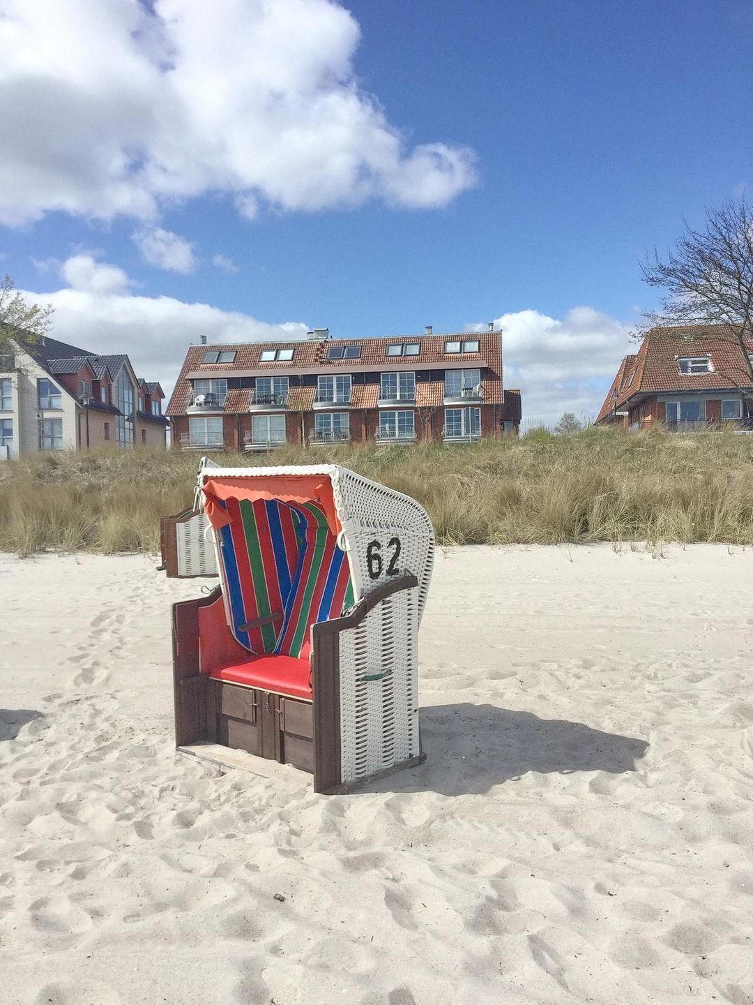 A beach chair with a colorful striped cushion and the number 62 on it, placed on a sandy beach with a row of houses in the background.