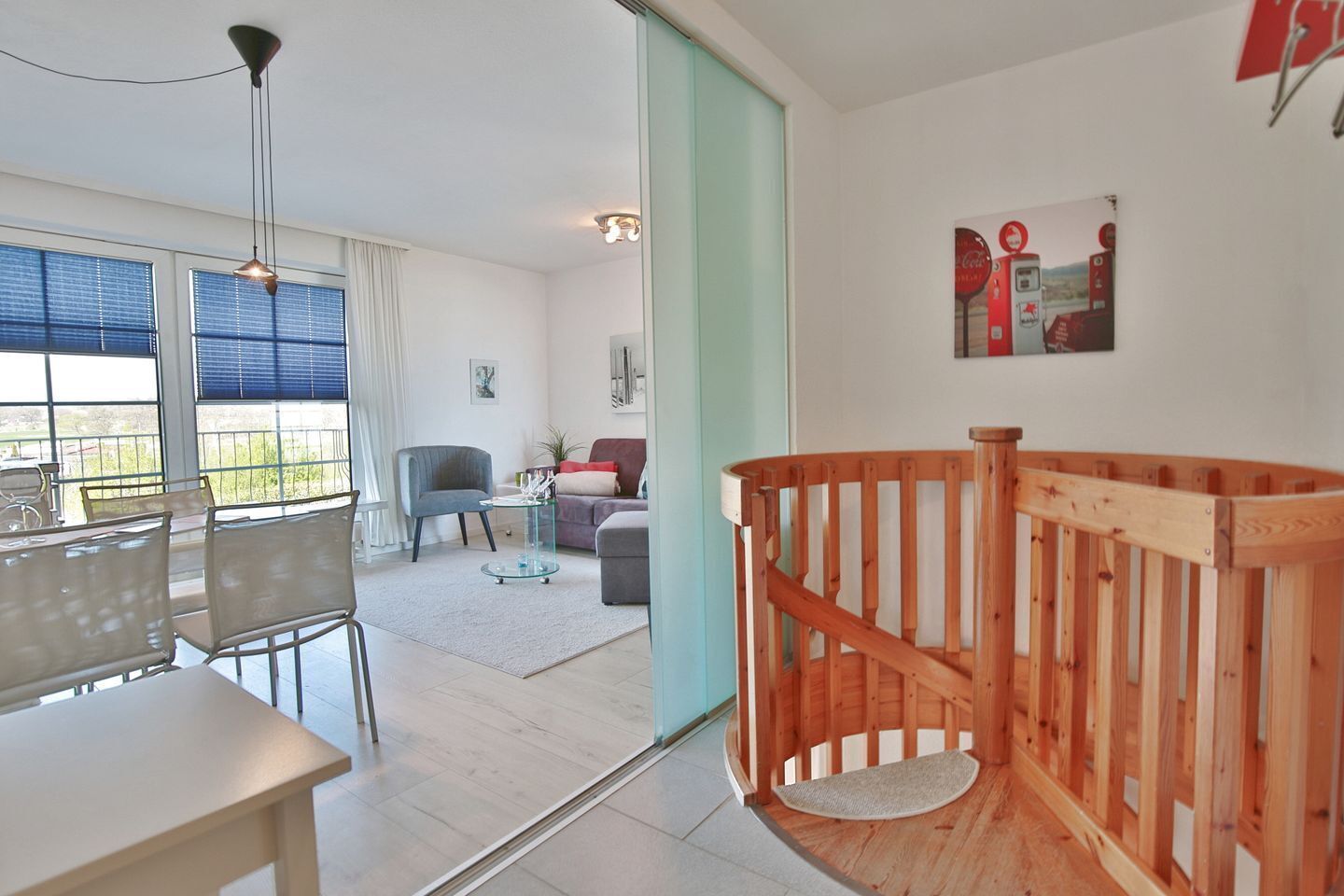 A modern living room with a wooden staircase, a dining area with chairs, and a large window with blinds.