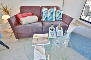 A living room with a brown leather sofa, a glass coffee table, and a blue armchair. There are two bottles of beer and a magazine on the table.