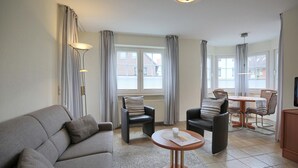 Een woonkamer met een bank, twee fauteuils, een salontafel en een eettafel met stoelen. Er hangen gordijnen voor de ramen, en een plafondlamp is zichtbaar.