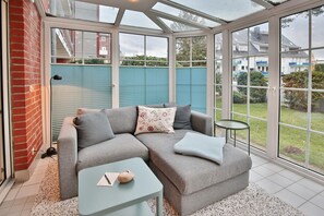 Une véranda avec un canapé gris, une petite table et une vue sur un jardin et des bâtiments.