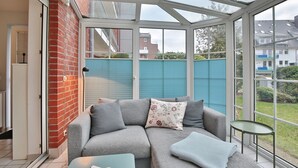 A sunroom with a glass ceiling, a grey sofa with cushions, a small round table, and a view of a blue fence and houses outside. - Windrose Appartement 1 - Haus Windrose (Scharbeutz)