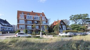 A multi-story residential building with balconies, a parking area with several cars, and surrounding greenery. - Windrose Appartement 1 - Haus Windrose (Scharbeutz)