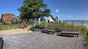 A wooden boardwalk with benches, a flag, a building with a red roof, and a grassy area with people walking. - Windrose Appartement 1 - Haus Windrose (Scharbeutz)