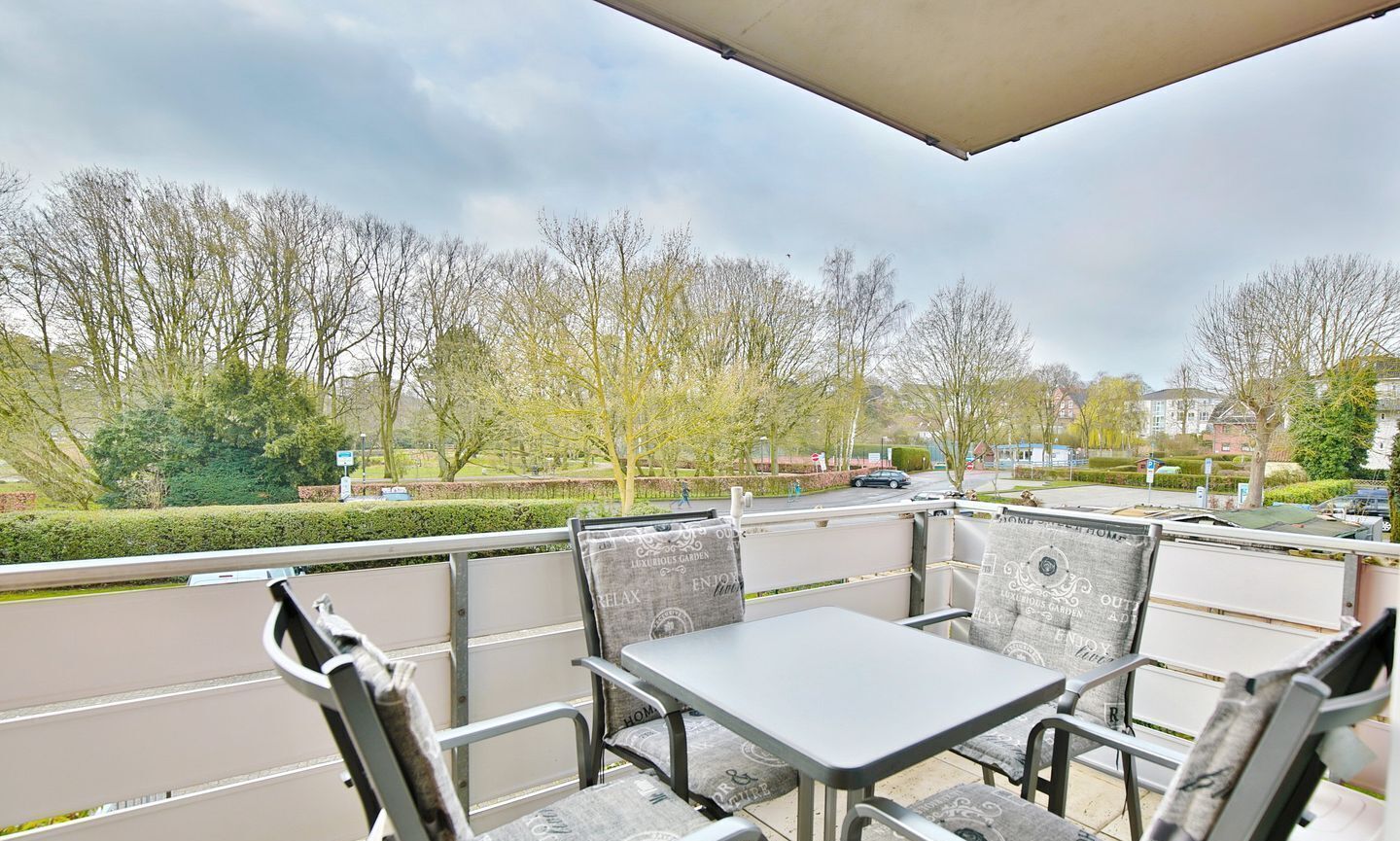 Un patio al aire libre con una mesa y sillas, vista a un parque con árboles y una calle, y un edificio al fondo.