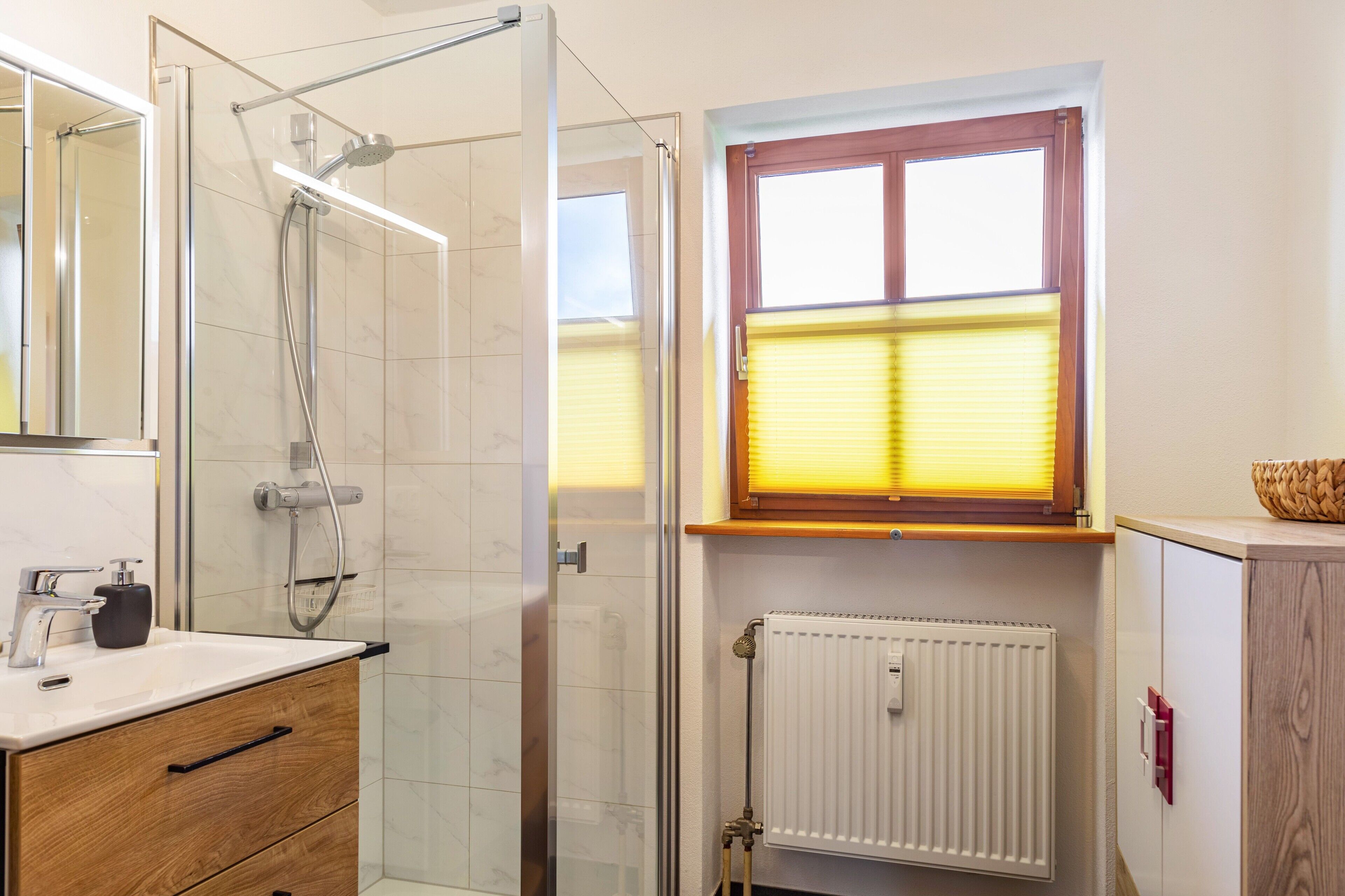 A bathroom with a glass shower enclosure, a white sink, wooden cabinet, and a window with blinds.