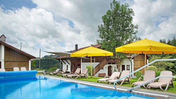A swimming pool with lounge chairs and yellow umbrellas, surrounded by wooden buildings and greenery. - Comfort apartment with pool, sauna and Wi-Fi (Hauzenberg)