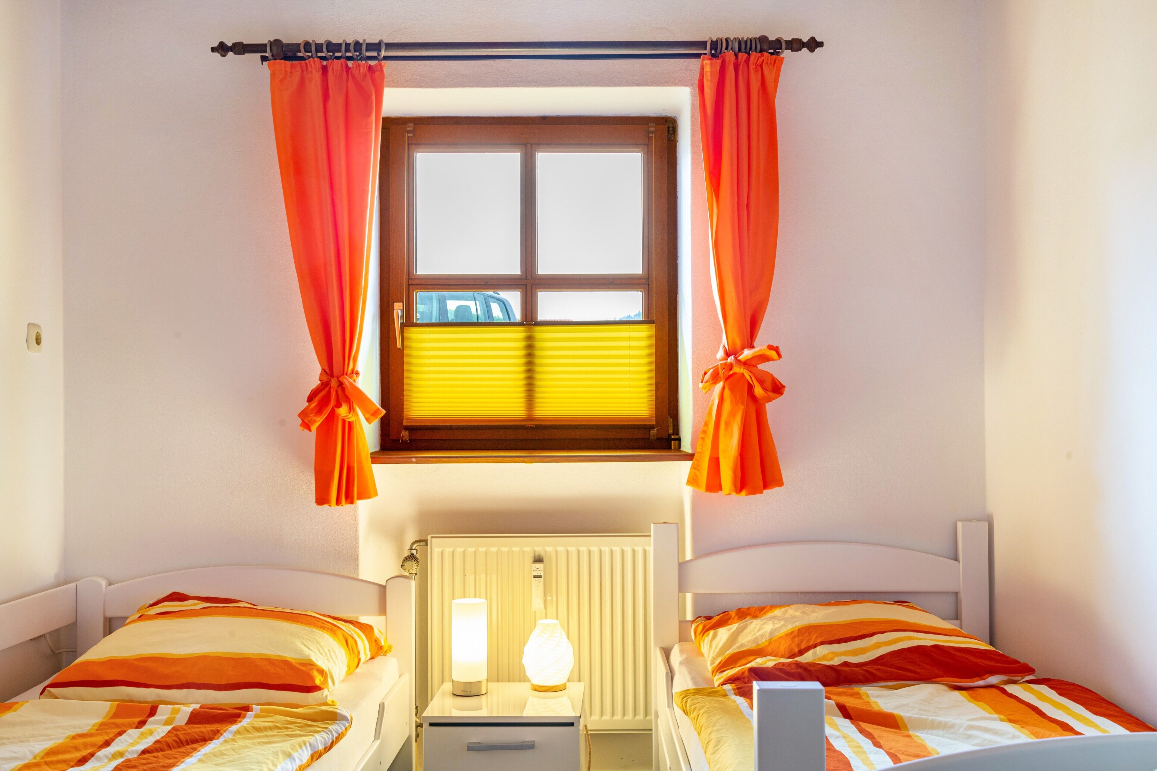 A bedroom with two beds, a window with curtains, a radiator, and bedside lamps.