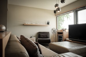 A modern living room with a sofa, a chair, a TV, a fan, and a shelf with glasses and bottles.