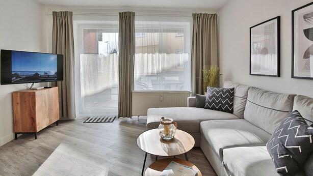 Un salón moderno con un sofá, una mesa de centro, un televisor sobre un mueble de madera y ventanas grandes con cortinas.
