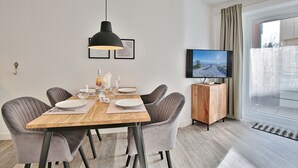 Un comedor moderno con una mesa de madera, seis sillas grises tapizadas, una lámpara colgante negra y un televisor sobre un mueble de madera.
