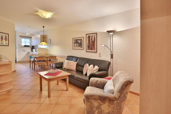 A living room with a sofa, armchair, and coffee table, a dining area with chairs and a table, and a kitchen in the background.
