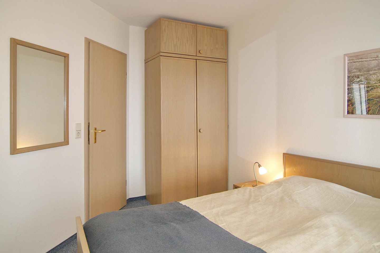 Een slaapkamer met een bed, een kledingkast, een nachtkastje met een lamp en een lijst aan de muur.