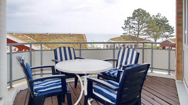 A covered outdoor seating area with a round table and striped chairs, overlooking a residential area with houses and a tree.