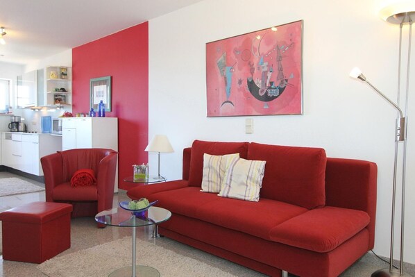 Ein rotes Sofa, ein Glastisch und ein rotes Sessel in einem modernen Wohnzimmer mit einer Küche im Hintergrund.