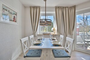 Un coin repas avec une table dressée pour quatre personnes, des chaises blanches, un luminaire suspendu et une fenêtre avec des rideaux.