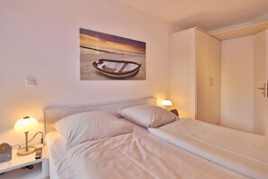 Ein Schlafzimmer mit Bett, Nachttischlampen, einem Nachttisch, einem Bild von einem Boot am Strand und einem Kleiderschrank.