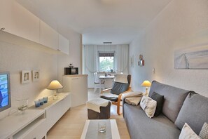Quarto de hotel moderno com sofá cinzento, televisão, secretária com luminária, cadeira e uma pequena cozinha equipada.