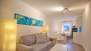 A modern living room with a grey sofa, patterned cushions, a white coffee table, and a large abstract painting. A dining area with a white table and chairs is visible in the background.