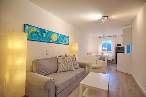 Una sala de estar moderna con un sofá gris, cojines estampados, una mesa de centro blanca y un cuadro abstracto de gran tamaño. Se observa, al fondo, una zona de comedor con una mesa y sillas blancas.