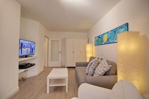 Un salon moderne avec un téléviseur à écran plat, une table basse blanche, un canapé gris agrémenté de coussins à motifs, une armoire blanche et un tableau accroché au mur.