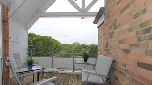 A covered outdoor balcony with a wooden deck, two metal chairs, a small table, and a potted plant.