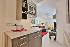 Una cocina moderna con microondas empotrado, tostadora y estantes. Se ve un área de comedor con un sofá gris y una alfombra roja al fondo.