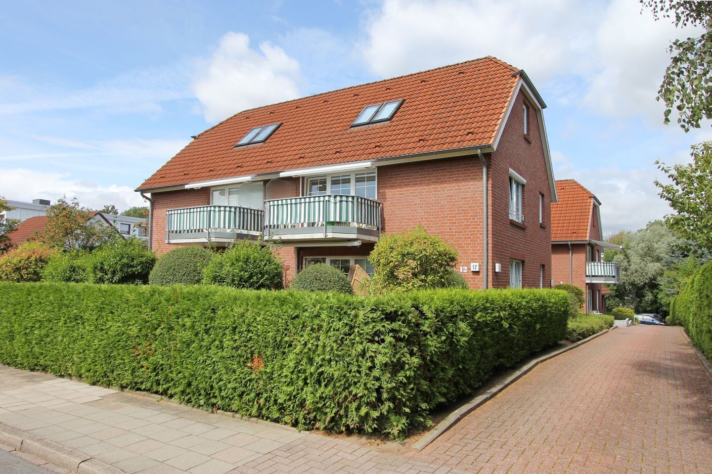 A two-story brick house with a red-tiled roof, a balcony with a white railing, and a well-maintained hedge in front.