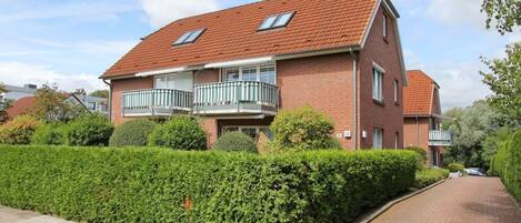 A two-story brick house with a red-tiled roof, a balcony with a white railing, and a well-maintained hedge in front.