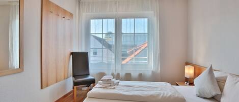 Habitación de hotel con cama, una silla, mesita de noche con lámpara y ventana con vista a un edificio.
