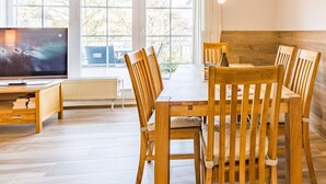 Un coin repas avec une table et des chaises en bois, un téléviseur posé sur un meuble, et de grandes fenêtres donnant sur la forêt.