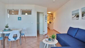 Una sala de estar moderna con un sofá azul, una zona de comedor con una mesa y sillas blancas, y una cocina al fondo.