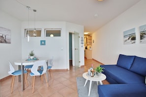 En moderne stue med en blå sofa, et spiseområde med hvide stole, og et køkken i baggrunden.