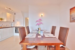 A dining area with a wooden table, wicker chairs, a vase of pink flowers, and a kitchen in the background.