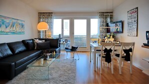 Una sala moderna con un sofá de cuero negro, una mesa de centro de vidrio y un comedor con sillas blancas y una pequeña mesa.