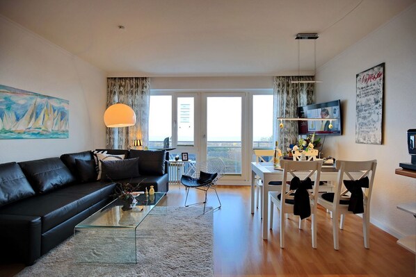 A modern living room with a black leather sofa, a glass coffee table, and a dining area with white chairs and a small table.