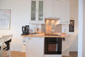 Cocina compacta con muebles blancos, horno empotrado, cafetera y un pequeño comedor con una silla.