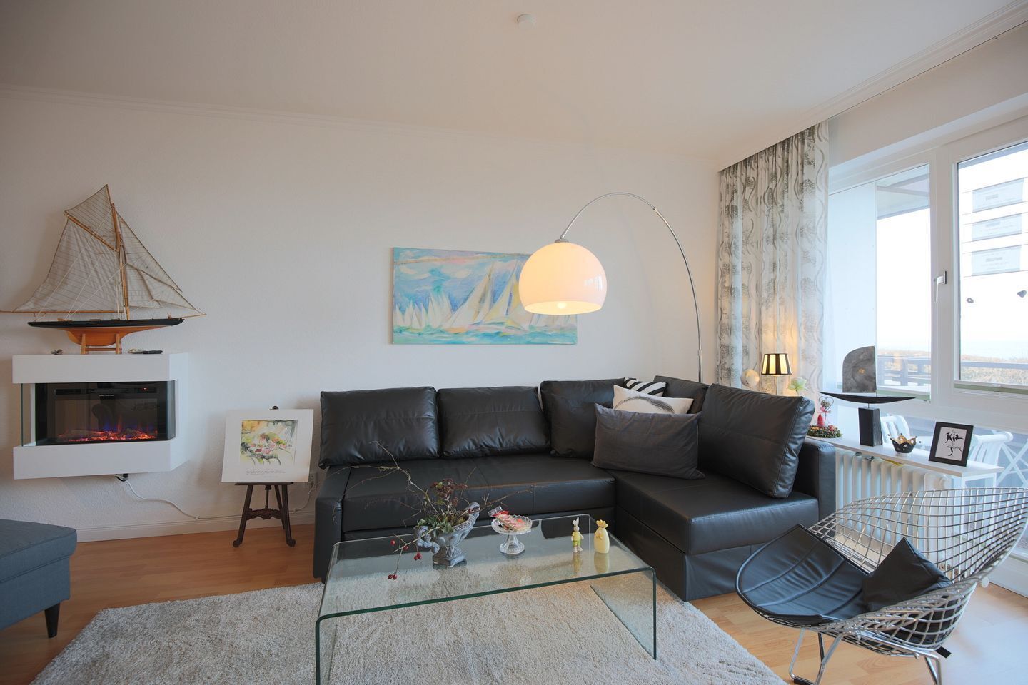 Een moderne woonkamer met een leren zwarte bank, een glazen salontafel, een klein model zeilboot en een schilderij van zeilboten.