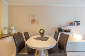 A dining area with a round table, four chairs, a coffee machine, a potted plant, a framed artwork, and a shelf with books and a speaker.