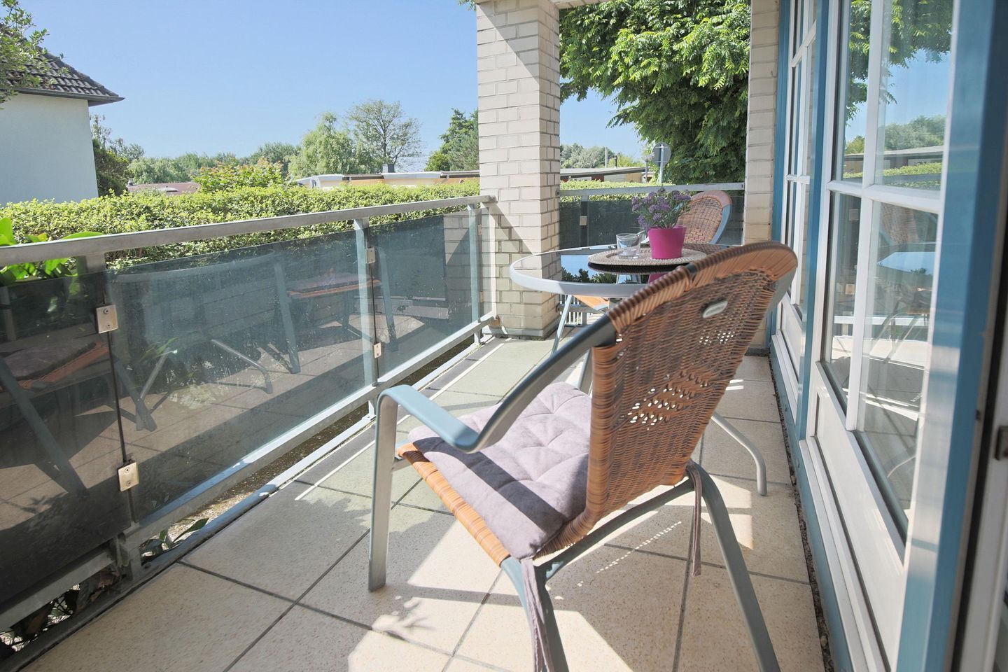 A balcony with a wicker chair, a small table with a pink vase, and a glass railing.