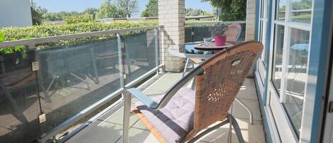 A balcony with a wicker chair, a small table with a pink vase, and a glass railing.