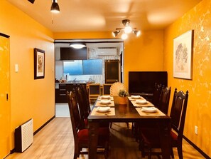 A dining area with a long table set for six, a television, and a kitchen in the background. - Art House  (東大阪市足代)