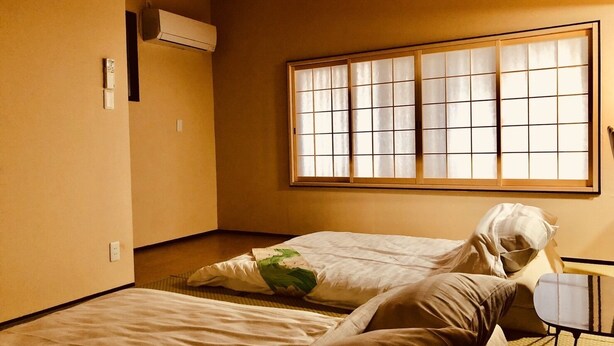 A room with two beds, a window with a grid pattern, an air conditioning unit, and a small table. - Art House  (東大阪市足代)