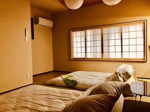 A room with two beds, a window with a grid pattern, an air conditioning unit, and a small table.