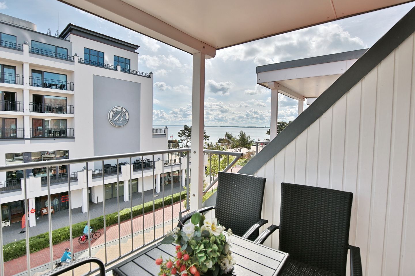 A balcony with wicker chairs and a table, overlooking a pedestrian street with shops and a view of the sea.