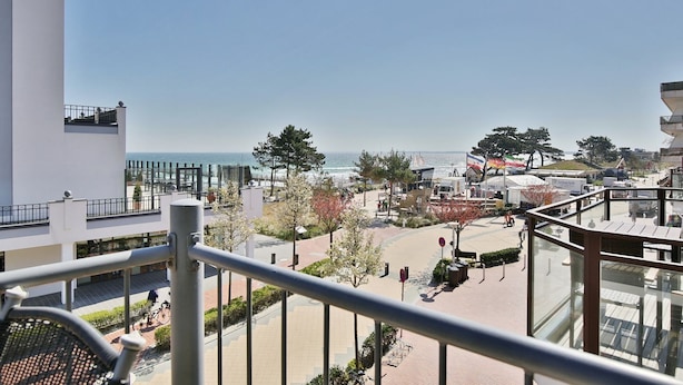 A balcony with a view of the beach, a parking area, and a building with a glass facade. - Seebrücke Appartement 2 - Seebrücke (Scharbeutz)