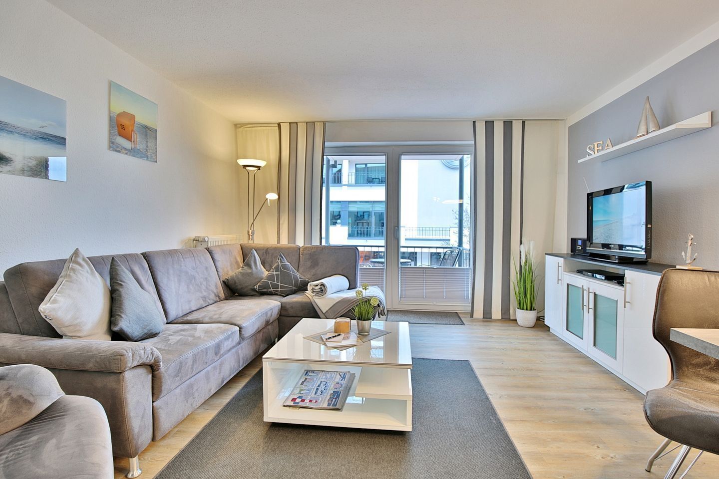 Un soggiorno moderno con un divano grigio, un tavolino da caffè con riviste, una TV su un mobile e un balcone con vista sugli edifici.